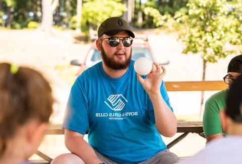 Man in Boys & Girls Club t-shirt holding a ball and talking to children