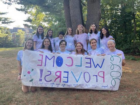 A group of students in tie-dye shirts holding a sign that reads "Welcome PrOVERS!"