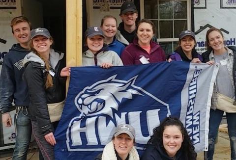 A group of students holding a UNH flag and standing in front of a house that they are building.