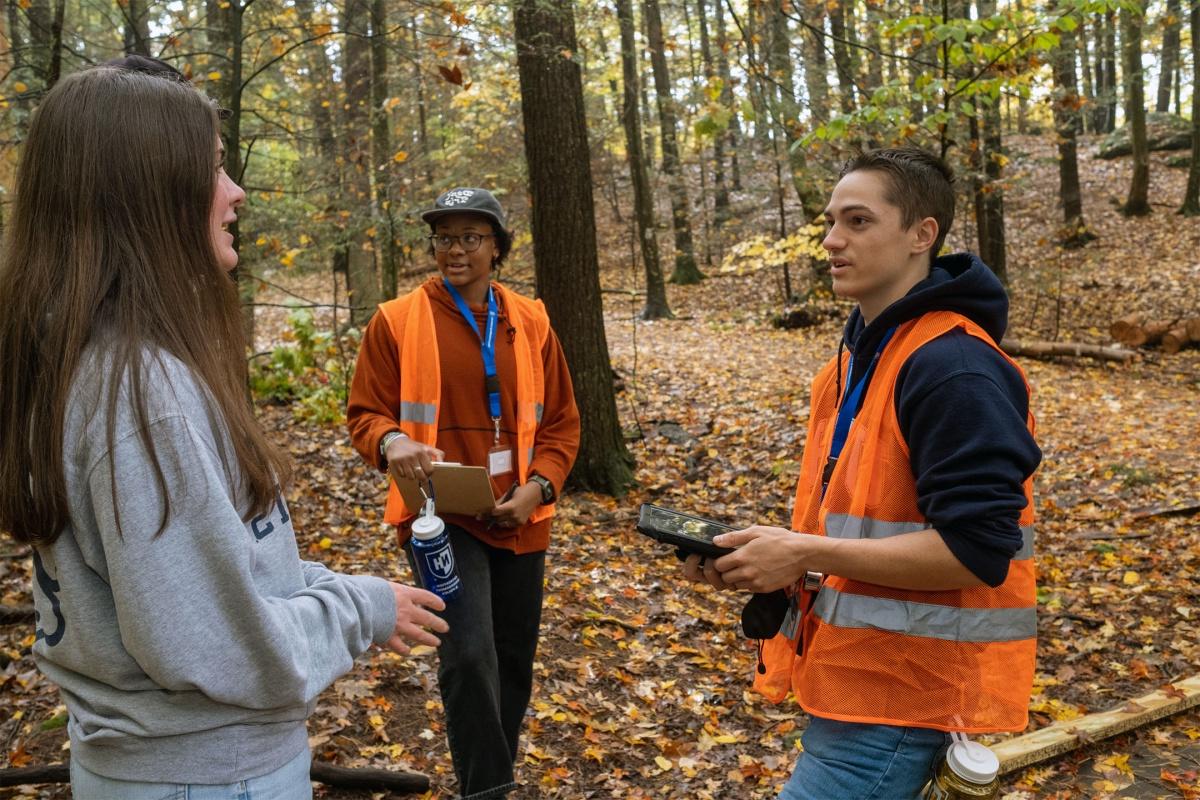 秋天，三个学生站在大学森林里. 两个学生穿着亮橙色的背心，假装在调查另一个学生. 第三个学生穿着一件灰色的运动衫.