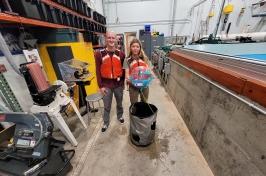 Two UNH student researchers hold their winning ocean renewable energy device in front of a wave tank. They are wearing red life jackets.